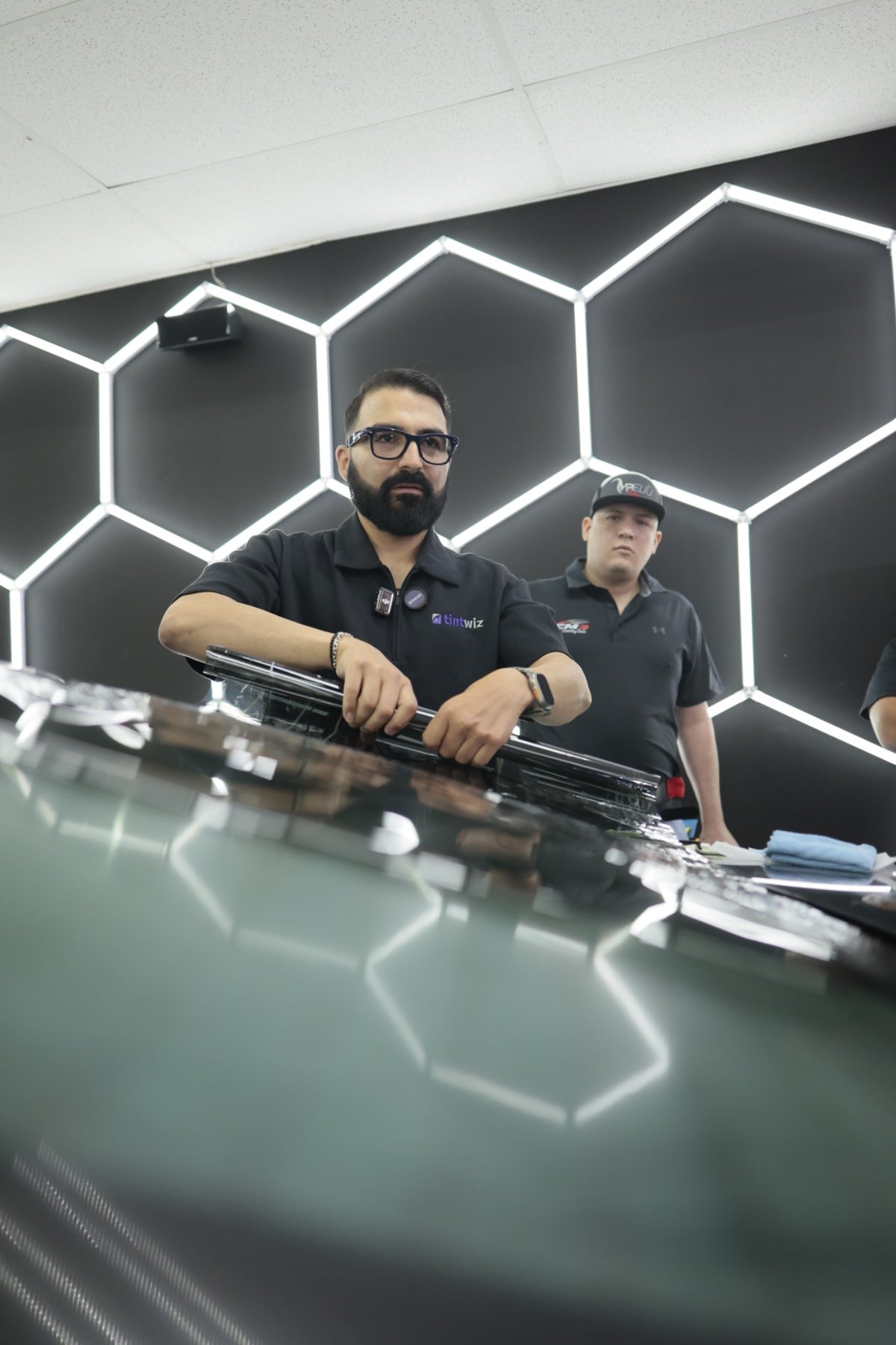 Marco demonstrates handling window tint film on a car while a student watches.